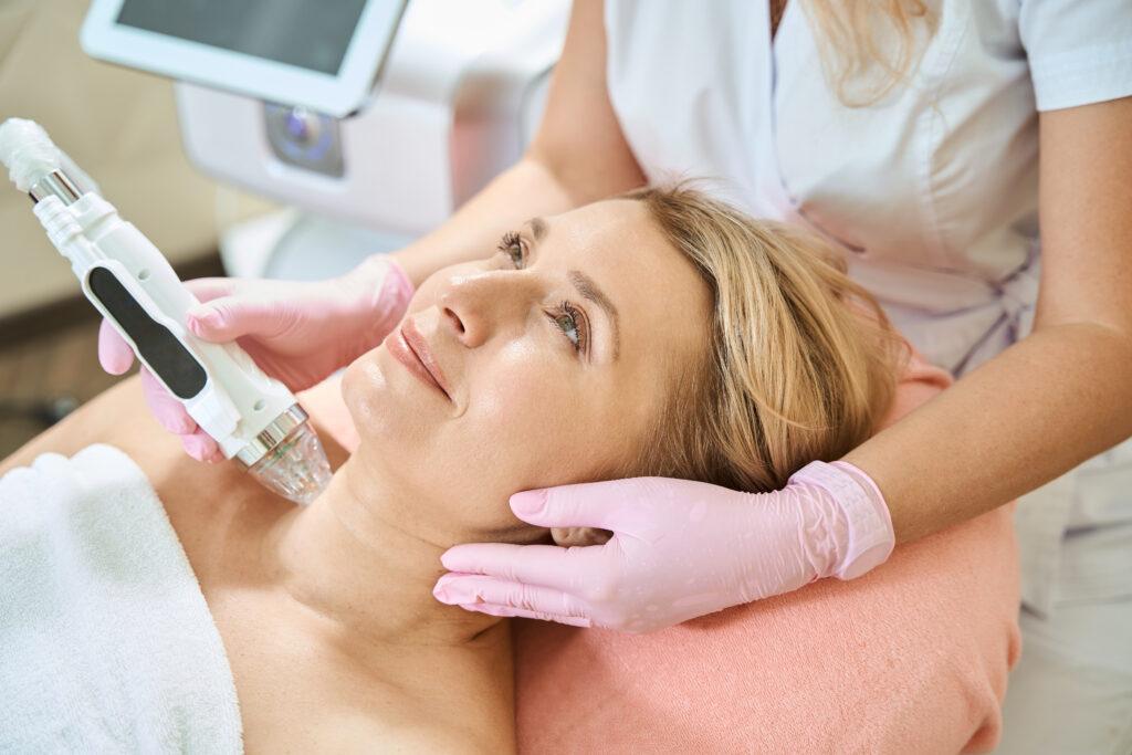 Female cosmetologist performing microneedle RF lifting and IPL photorejuvenation on a smiling woman’s neck, highlighting professional body skin care and rejuvenation.